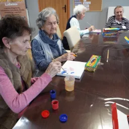 Grupa starszych kobiet siedzi przy stole i maluje obrazy na płótnie, korzystając z farb i pędzli. Na stole widoczne są pojemniki z farbami, pędzle i pudełka z farbami. W tle widoczny plakat...