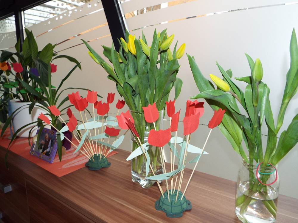 Kompozycja z żywych żółtych tulipanów i papierowych czerwonych tulipanów w szklanych wazonach, obok zdjęcie w ramce i doniczka z zieloną rośliną na drewnianej komodzie.