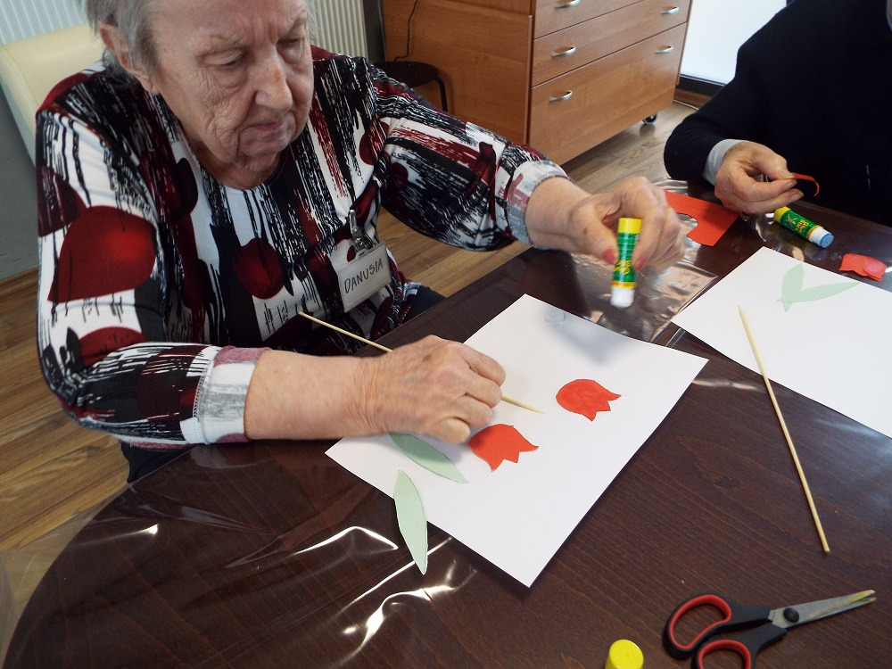Starsza kobieta z identyfikatorem Danusia przykleja czerwone elementy tulipanów do kartki, używając kleju w sztyfcie; obok leżą zielone liście i patyczki do szaszłyków.