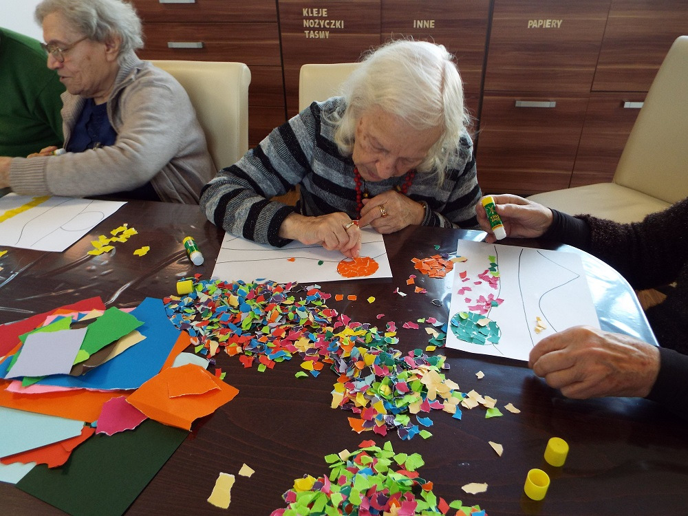 Seniorzy podczas zajęć plastycznych, wyklejanie obrazków z kolorowych skrawków papieru, widoczne kleje w sztyfcie, różnokolorowe resztki papieru na stole.