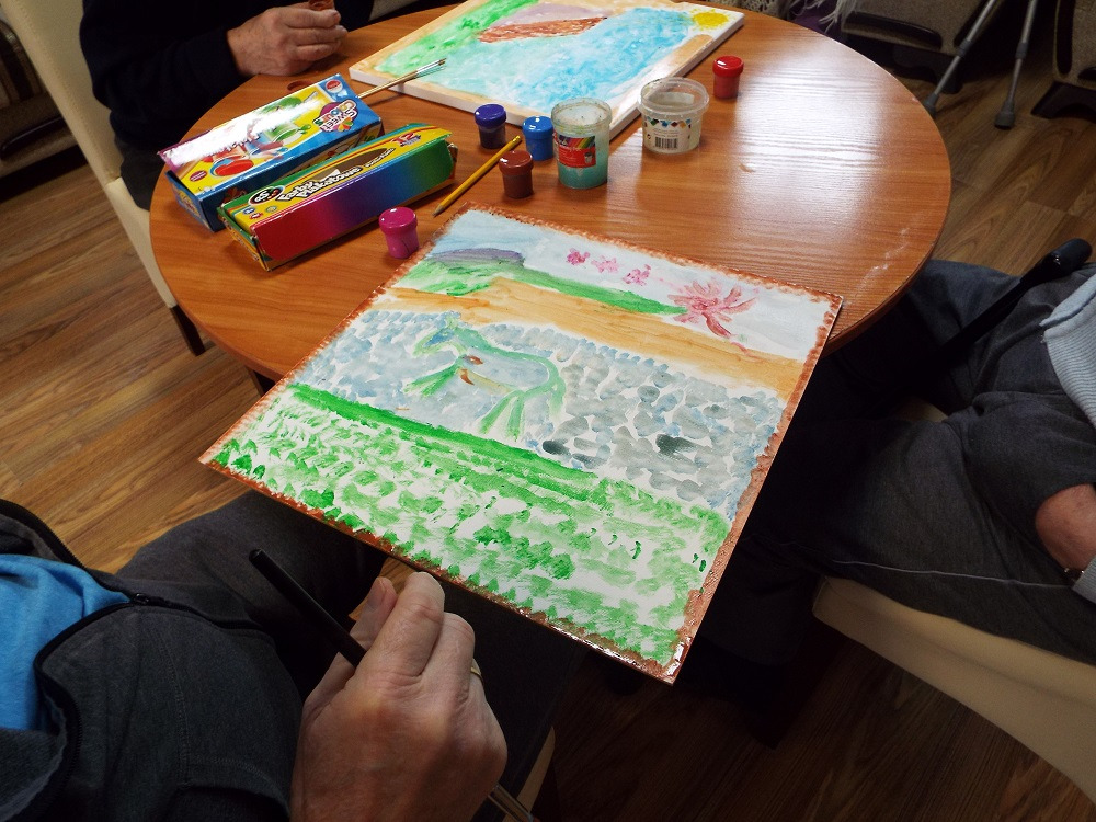 Osoba trzyma obraz z namalowanym pejzażem, farby i pędzle leżą na okrągłym stole, w tle druga osoba maluje inny obraz.