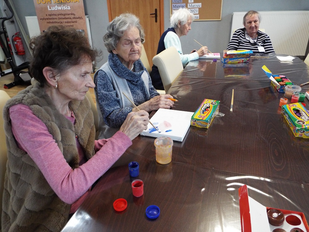 Grupa starszych kobiet siedzi przy stole i maluje obrazy na płótnie, korzystając z farb i pędzli. Na stole widoczne są pojemniki z farbami, pędzle i pudełka z farbami. W tle widoczny plakat...