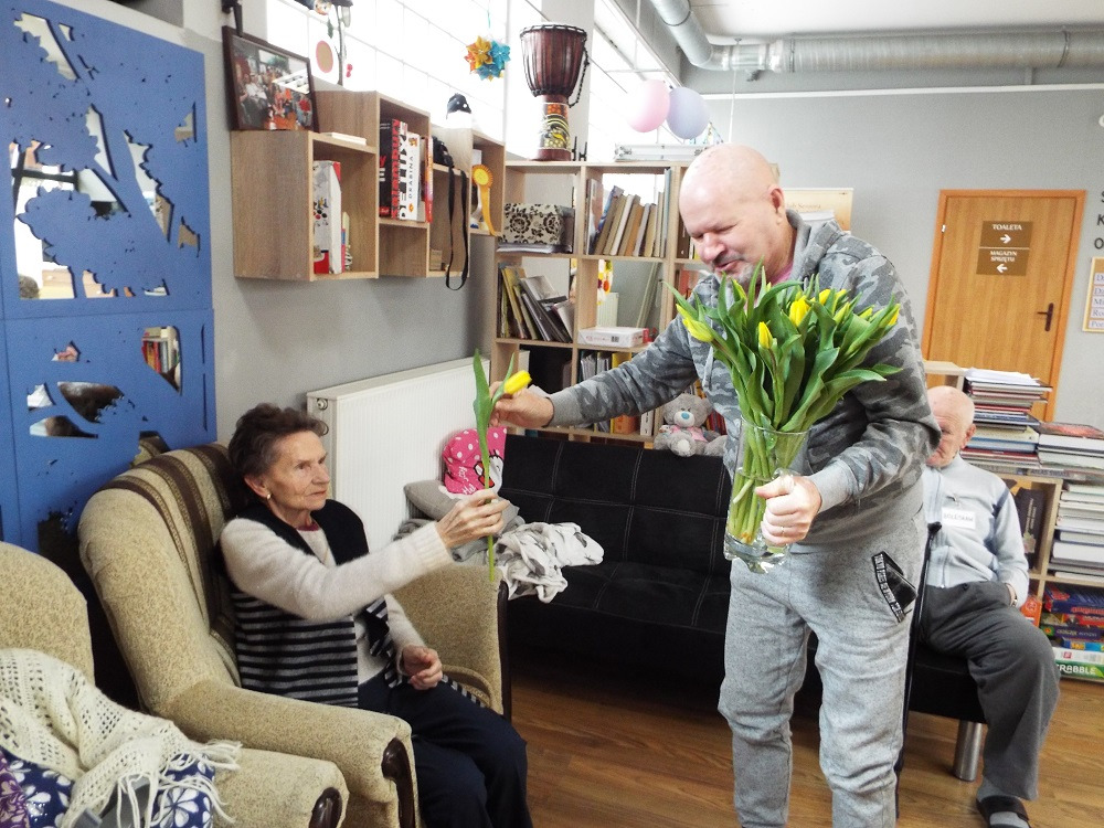 Mężczyzna wręcza bukiet żółtych tulipanów starszej kobiecie siedzącej w fotelu w pokoju z regałami książek.