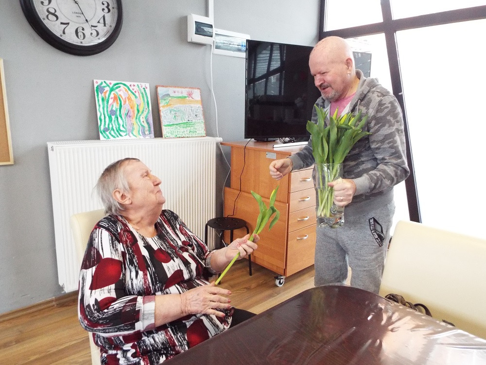 Opiekun wręcza tulipany starszej kobiecie siedzącej przy stole w pokoju z dekoracjami dziecięcymi na ścianie.