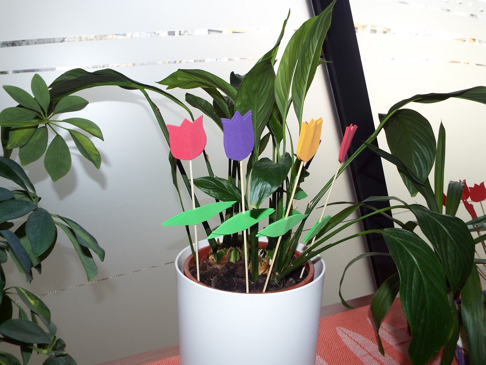 Donica z rośliną ozdobiona kolorowymi, papierowymi tulipanami na patyczkach, ustawiona w pomieszczeniu z widocznym oknem.