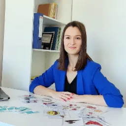 Kobieta w niebieskiej marynarce siedzi przy biurku z laptopem i kartami obrazkowymi, uśmiechając się.