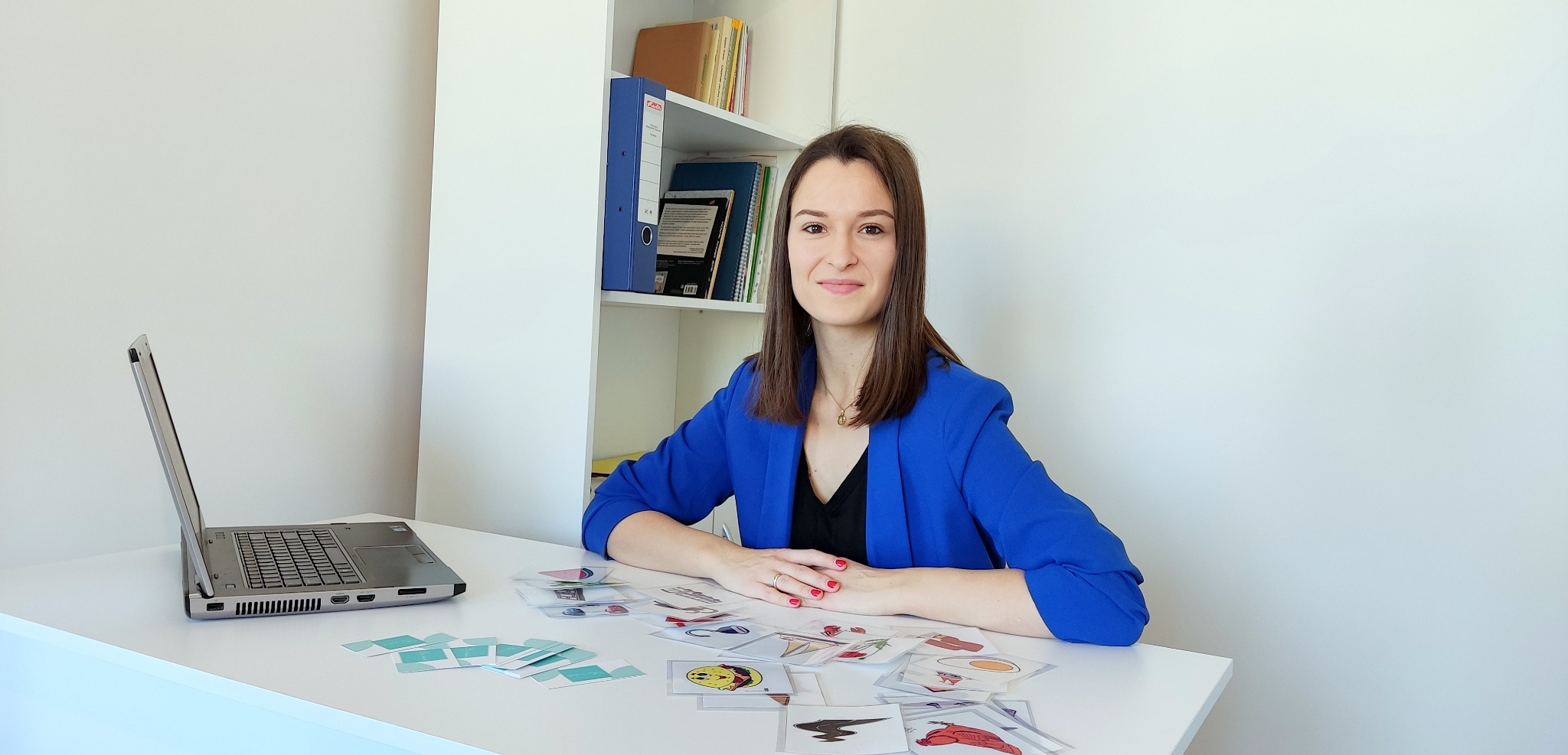 Kobieta w niebieskiej marynarce siedzi przy biurku z laptopem i kartami obrazkowymi, uśmiechając się.