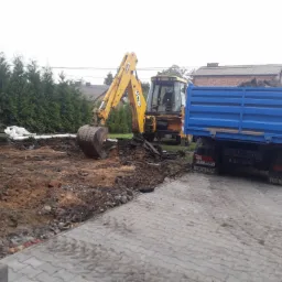 Żółta koparka JCB i niebieska wywrotka Renault na placu budowy, widoczne świeżo wykopana ziemia i kostka brukowa, w tle zieleń i budynek.