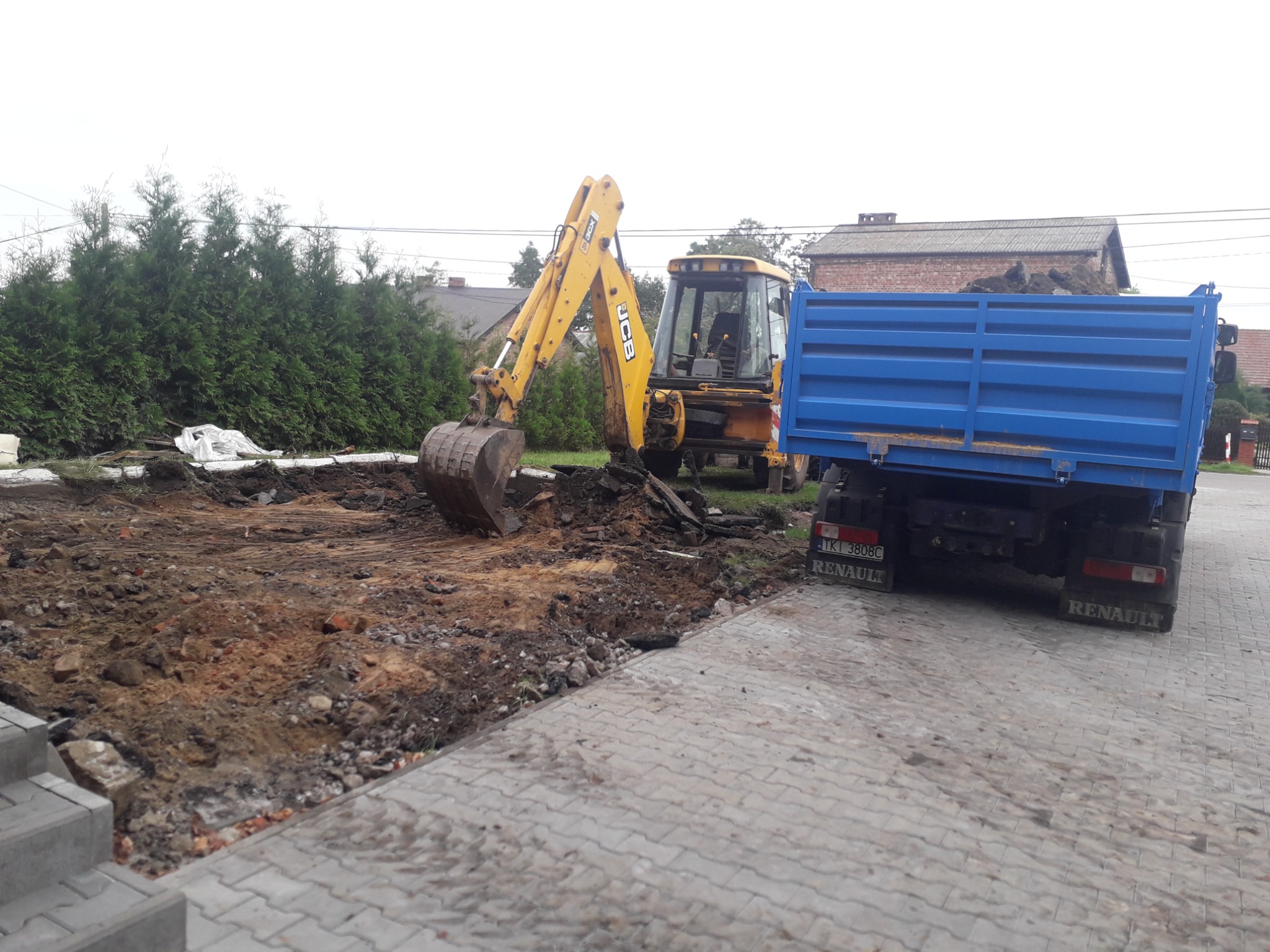 Żółta koparka JCB i niebieska wywrotka Renault na placu budowy, widoczne świeżo wykopana ziemia i kostka brukowa, w tle zieleń i budynek.