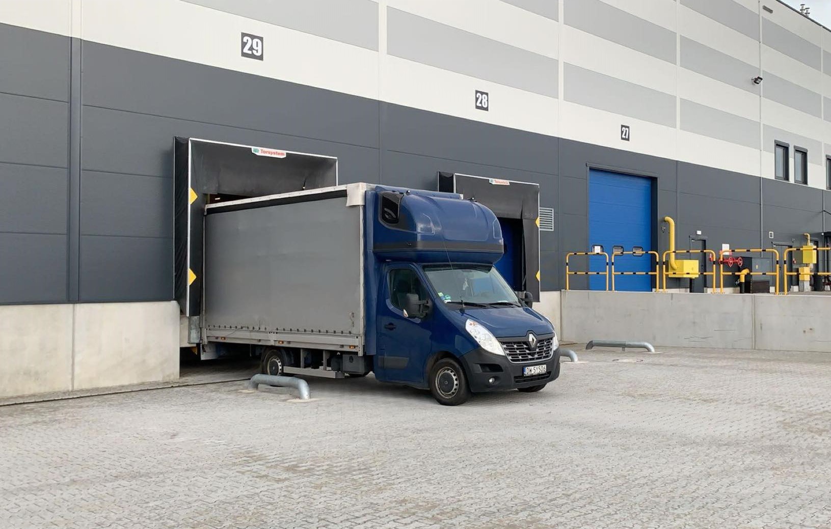 Niebieski bus dostawczy Renault Master z szarą zabudową wjeżdża do rampy załadunkowej magazynu o numerze 29, widoczne żółte oznaczenia na rampie.