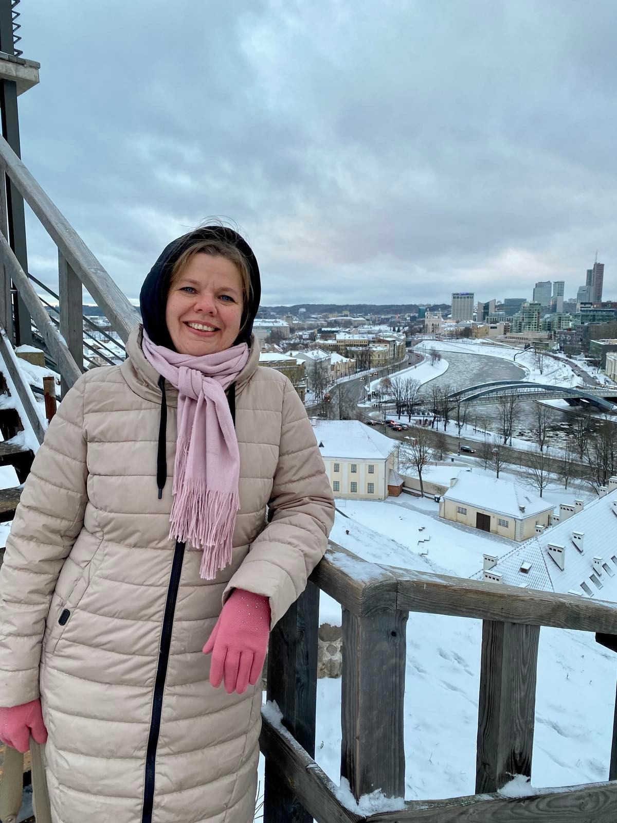 Uśmiechnięta kobieta w beżowej kurtce i różowym szaliku oraz rękawiczkach pozuje na drewnianym tarasie widokowym z panoramą zasypanego śniegiem miasta w tle.