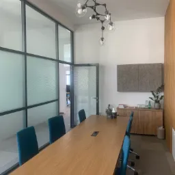 Nowoczesna sala konferencyjna z dużym drewnianym stołem, turkusowymi krzesłami, przeszkloną ścianą z falistym wzorem i modernistyczną lampą sufitową.