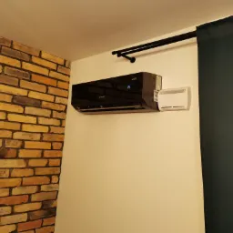 Czarny klimatyzator ścienny Rotenso z wyświetlaczem temperatury 16 stopni, zamontowany na białej ścianie obok dekoracyjnej ściany z cegły i zasłony w kolorze ciemnej zieleni.