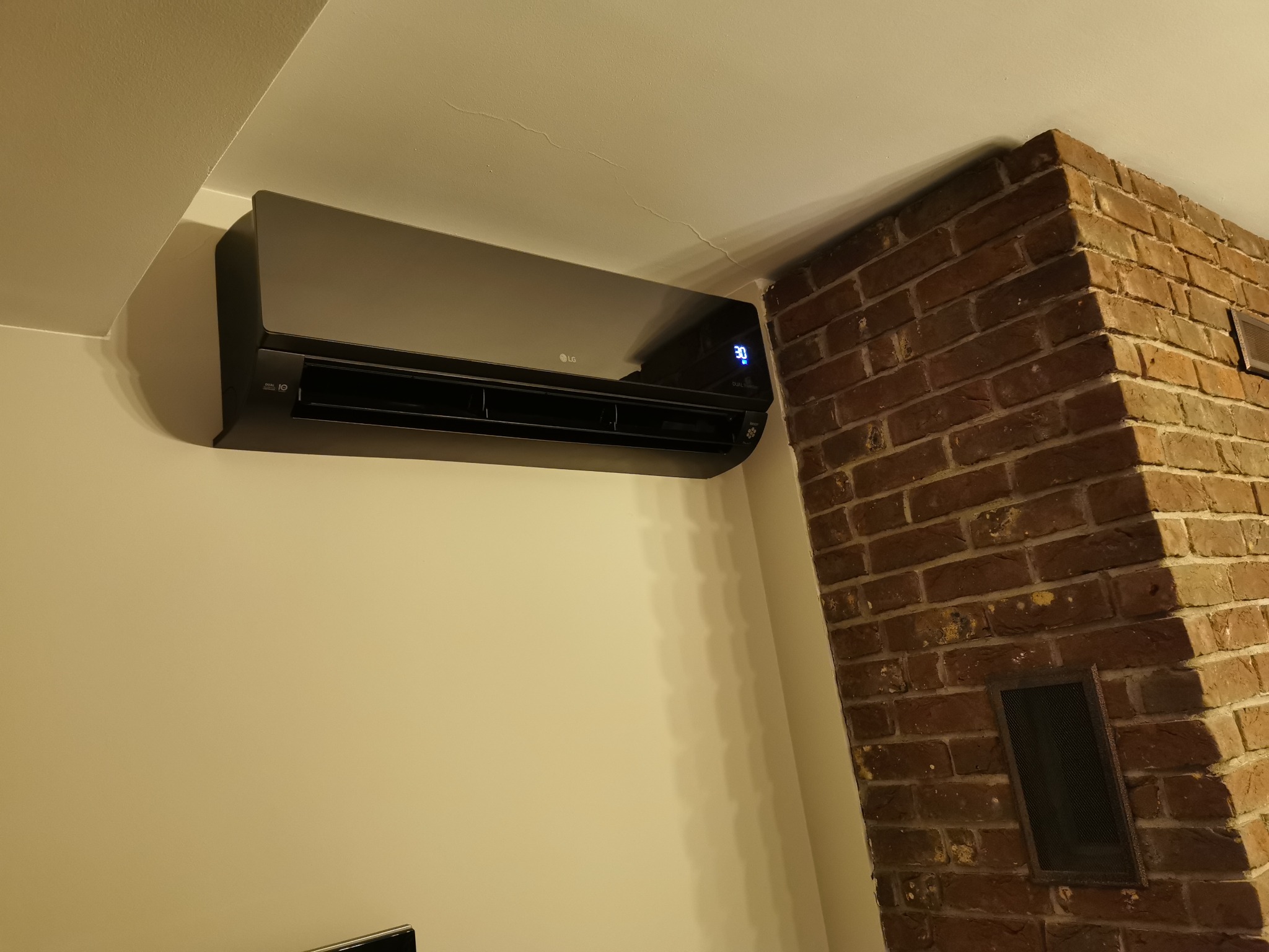 Czarny klimatyzator LG zamontowany na ścianie w pomieszczeniu z ceglanym kominem, wyświetlający temperaturę 30 stopni Celsjusza.