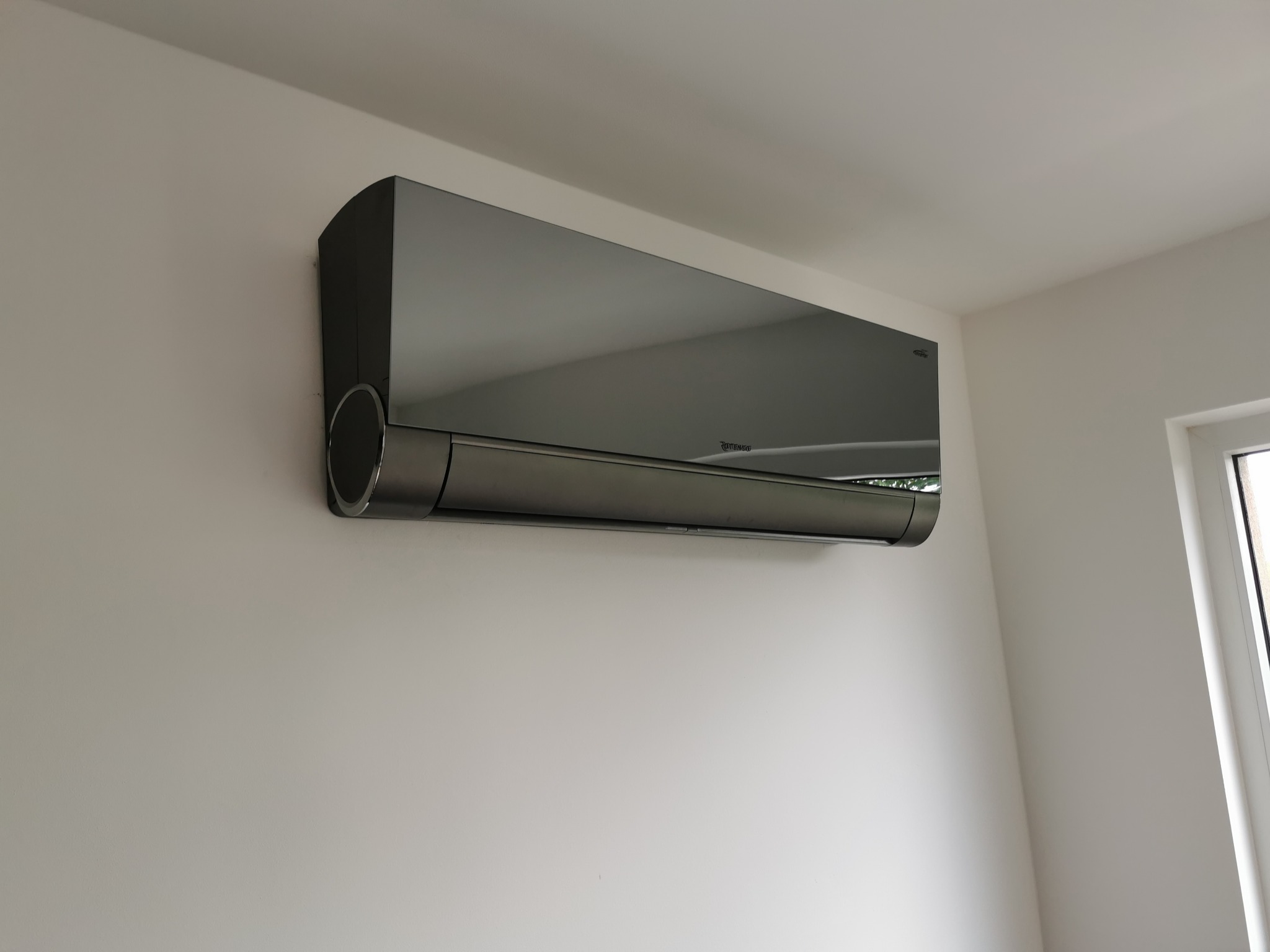 Czarny klimatyzator ścienny zamontowany w rogu pokoju z białymi ścianami, widoczny fragment okna.