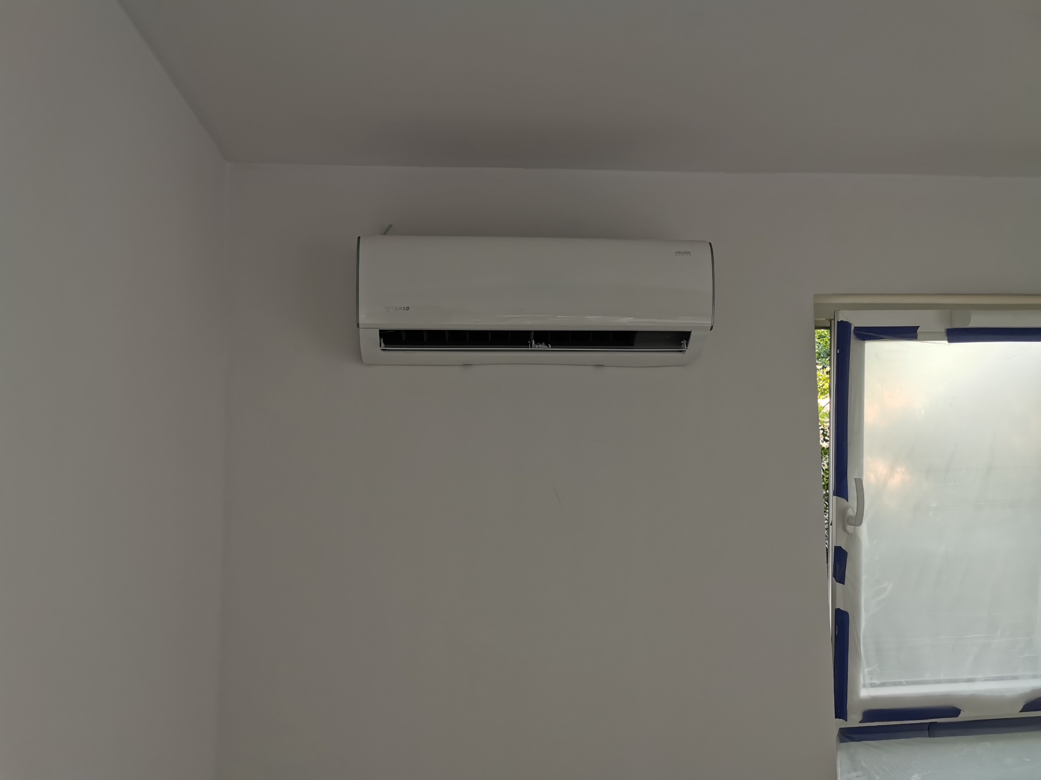 Biały klimatyzator zamontowany na ścianie w pokoju, okno zabezpieczone folią malarską.