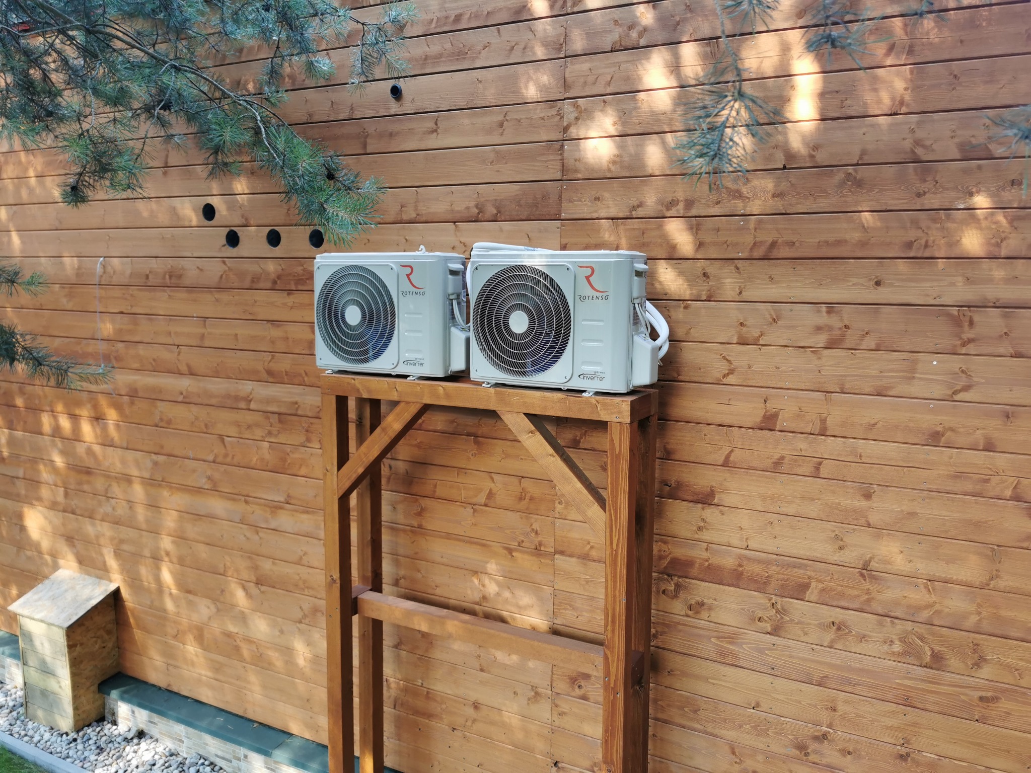 Dwa nowe klimatyzatory marki Rotenso ustawione na drewnianym stelażu przed drewnianą ścianą w Warszawie, widoczne także nawiercone otwory pod instalację.