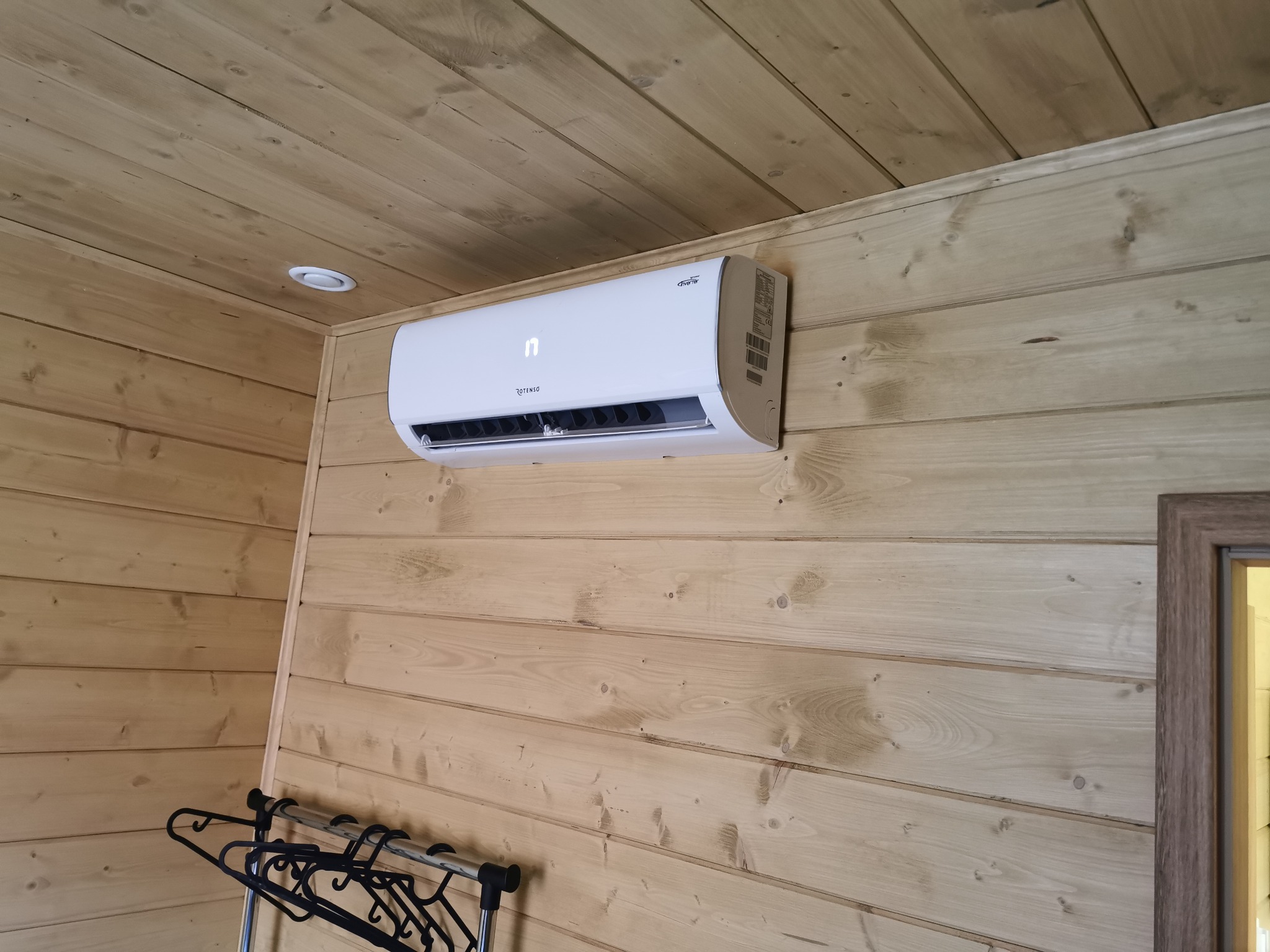 Biały klimatyzator marki Rotenso zamontowany na drewnianej ścianie w domu z drewnianą boazerią, widoczne wieszaki na ubrania w rogu pomieszczenia.