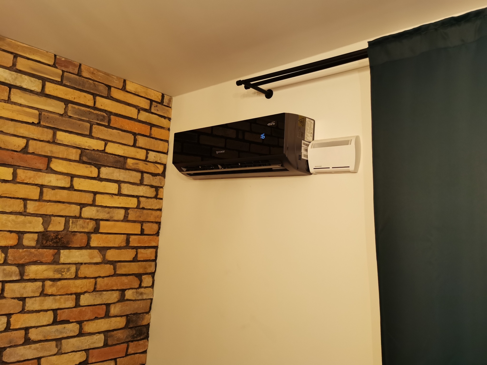 Czarny klimatyzator ścienny Rotenso z wyświetlaczem temperatury 16 stopni, zamontowany na białej ścianie obok dekoracyjnej ściany z cegły i zasłony w kolorze ciemnej zieleni.