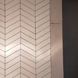 Układanie białych płytek w jodełkę na ścianie z widocznymi krzyżykami dystansowymi, obok fragment ściany z płytami gipsowo-kartonowymi.