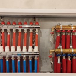 Hydro-Now - Rozdzielacz ogrzewania podłogowego w skrzynce, z rurami w kolorze czerwonym i niebieskim, z widocznymi zaworami i numeracją sekcji.