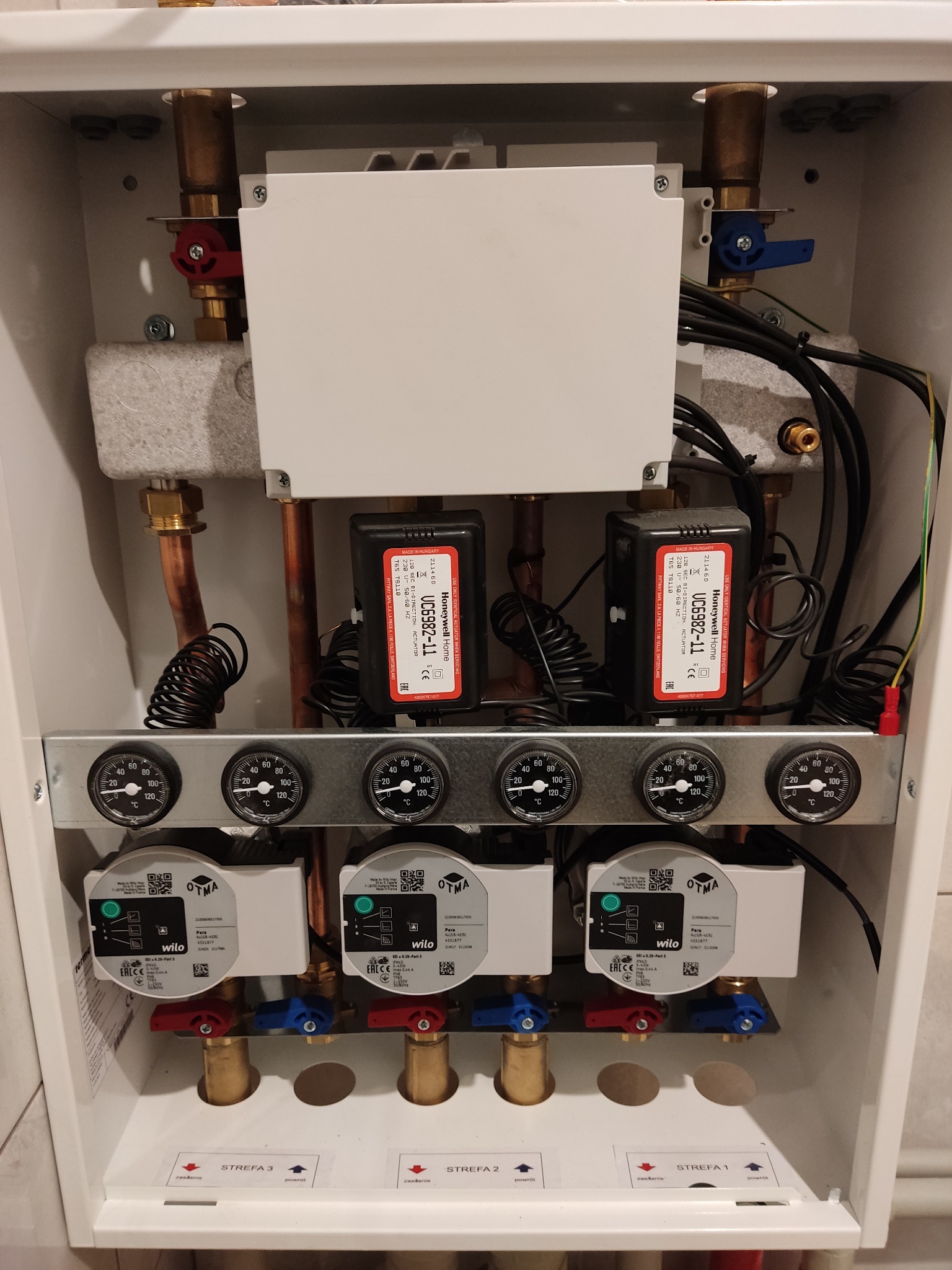 Rozdzielacz strefowy ogrzewania podłogowego w szafce, widoczne pompy obiegowe Wilo, termometry, zawory kulowe z czerwonymi i niebieskimi uchwytami oraz sterowniki Honeywell Home.
