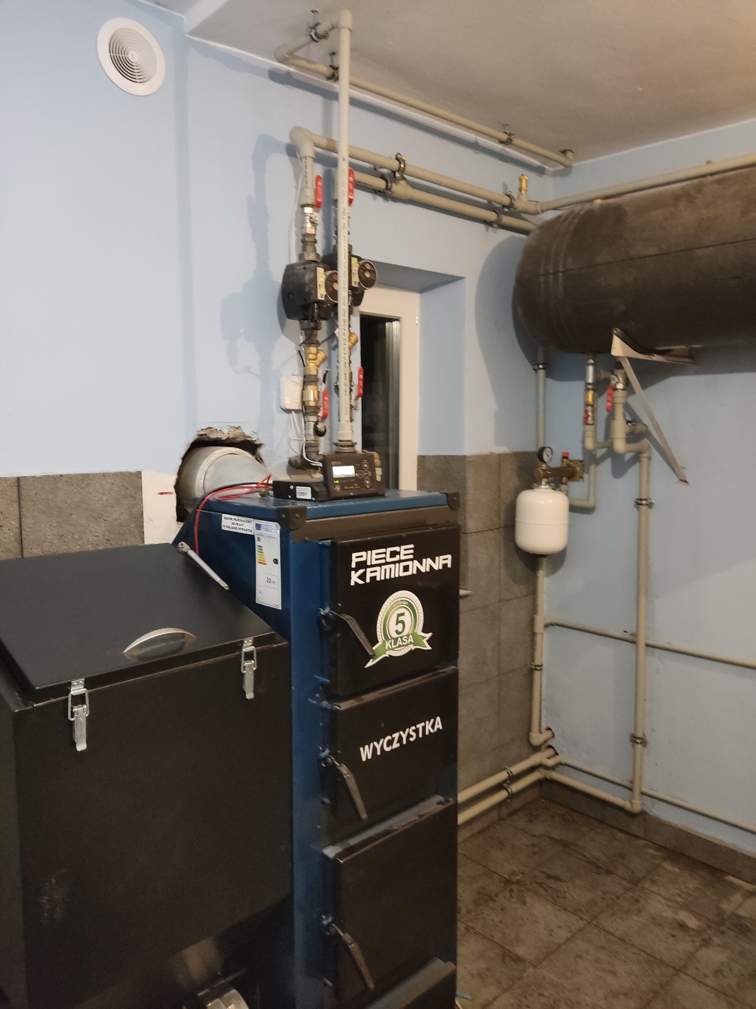 Kocioł na paliwo stałe z podajnikiem, widoczne rury instalacji grzewczej, zbiornik akumulacyjny i elementy armatury w kotłowni o ścianach wykończonych częściowo płytkami.