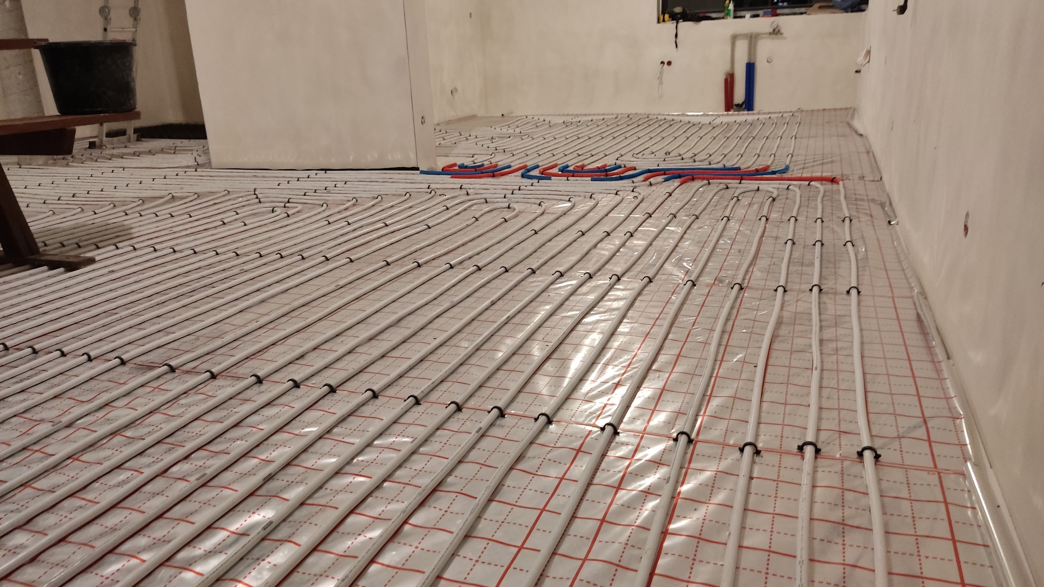 Montaż ogrzewania podłogowego: białe rurki grzewcze mocowane czarnymi uchwytami do folii z czerwoną siatką w surowym wnętrzu.