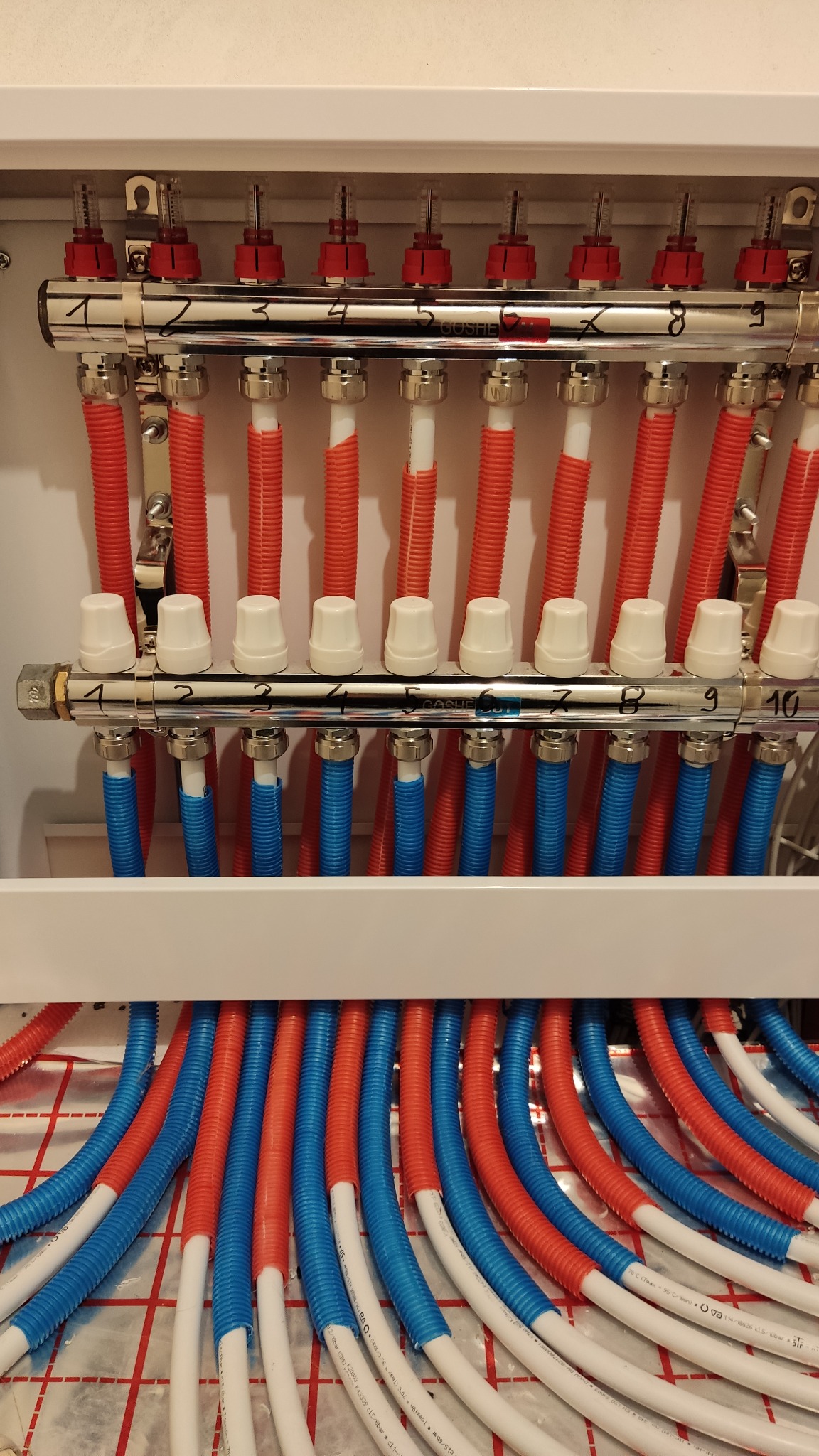 Rozdzielacz ogrzewania podłogowego z rurami w kolorach czerwonym i niebieskim, zamontowany w szafce, widoczne numery na belkach rozdzielacza.