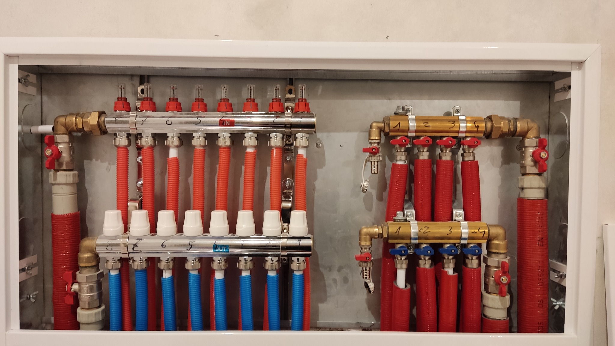 Rozdzielacz ogrzewania podłogowego w skrzynce, z rurami w kolorze czerwonym i niebieskim, z widocznymi zaworami i numeracją sekcji.