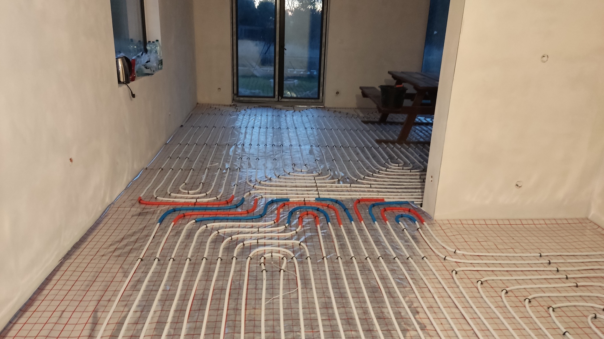 Rozłożone rury ogrzewania podłogowego w nowym budynku, widoczne białe, czerwone i niebieskie rury na siatce, przygotowane do zalania wylewką.