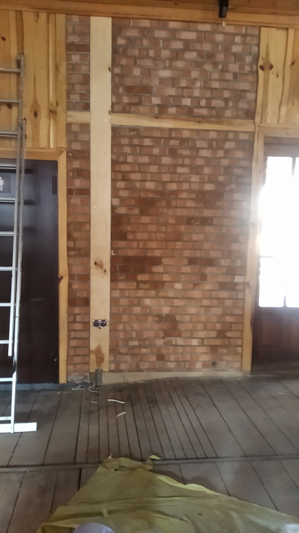 Wnętrze w trakcie budowy z odsłoniętą ceglaną ścianą, drewnianą boazerią, widoczną instalacją elektryczną i fragmentem drabiny opartą o ścianę.