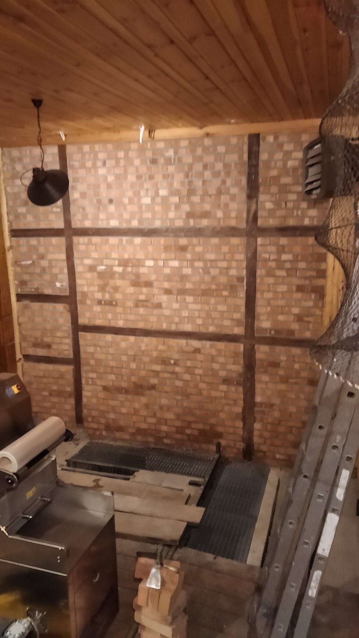 Wnętrze w trakcie budowy z surową ceglaną ścianą, drewnianą konstrukcją, widocznymi kratkami wentylacyjnymi w podłodze, sprzętem budowlanym i drabiną opartą o ścianę.