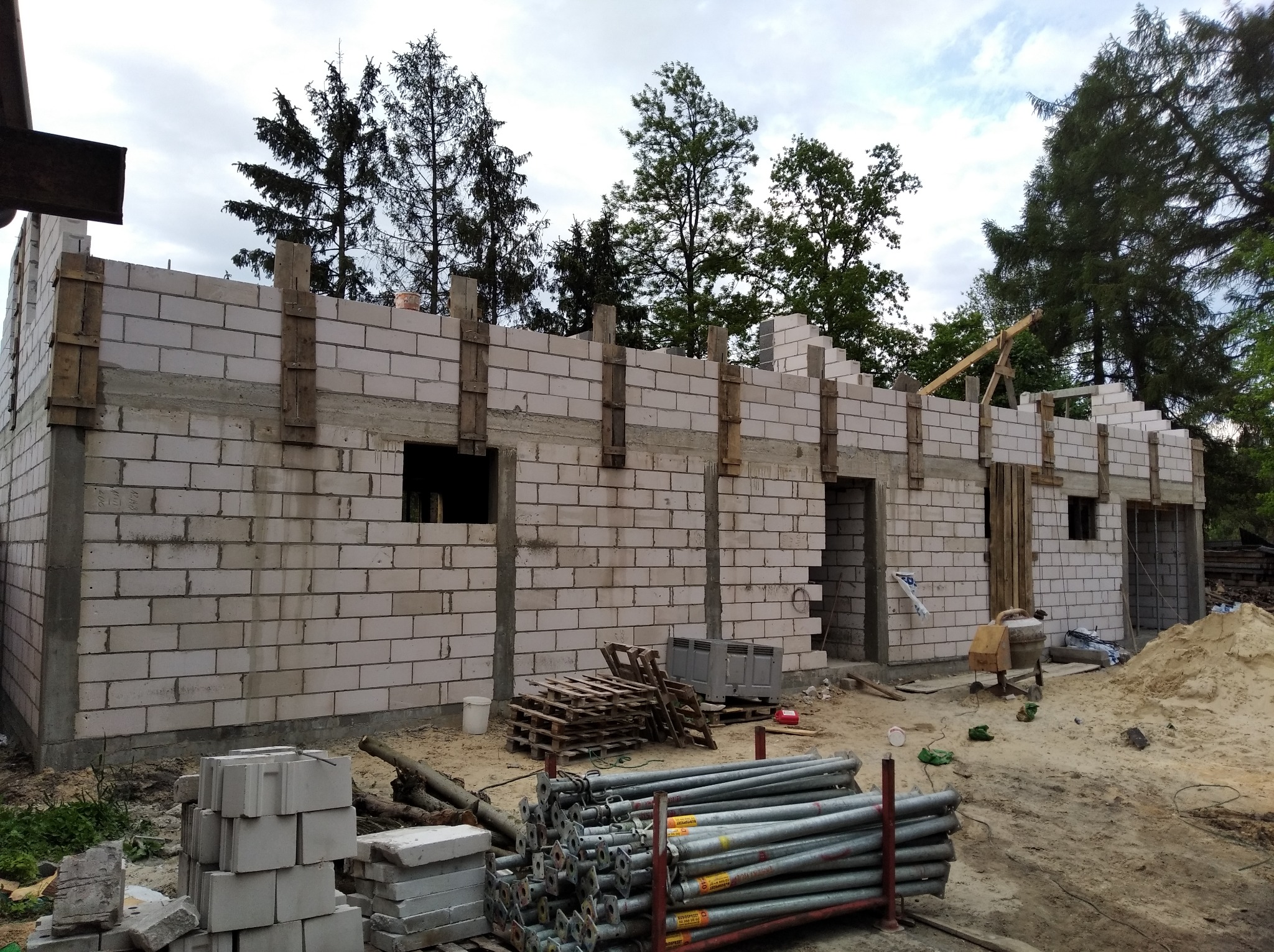 Surowy stan domu z białych bloczków, widoczne drewniane szalunki i podpory, stosy materiałów budowlanych i piasku na placu budowy.