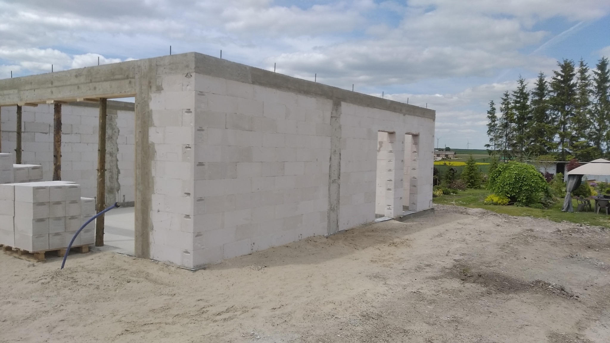 Surowy stan budynku z białych bloczków, widoczne otwory okienne i drzwiowe, betonowy wieniec, na palecie białe bloczki, teren wokół w stanie surowym, w tle zieleń i błękitne niebo.