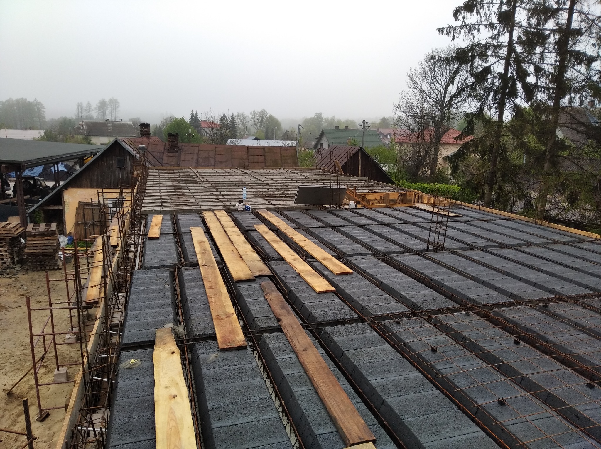 Wznoszenie stropu Teriva na budowie domu jednorodzinnego, widok z góry na szare betonowe pustaki i drewniane deski, w tle zabudowa mieszkalna i drzewa w lekkiej mgle.