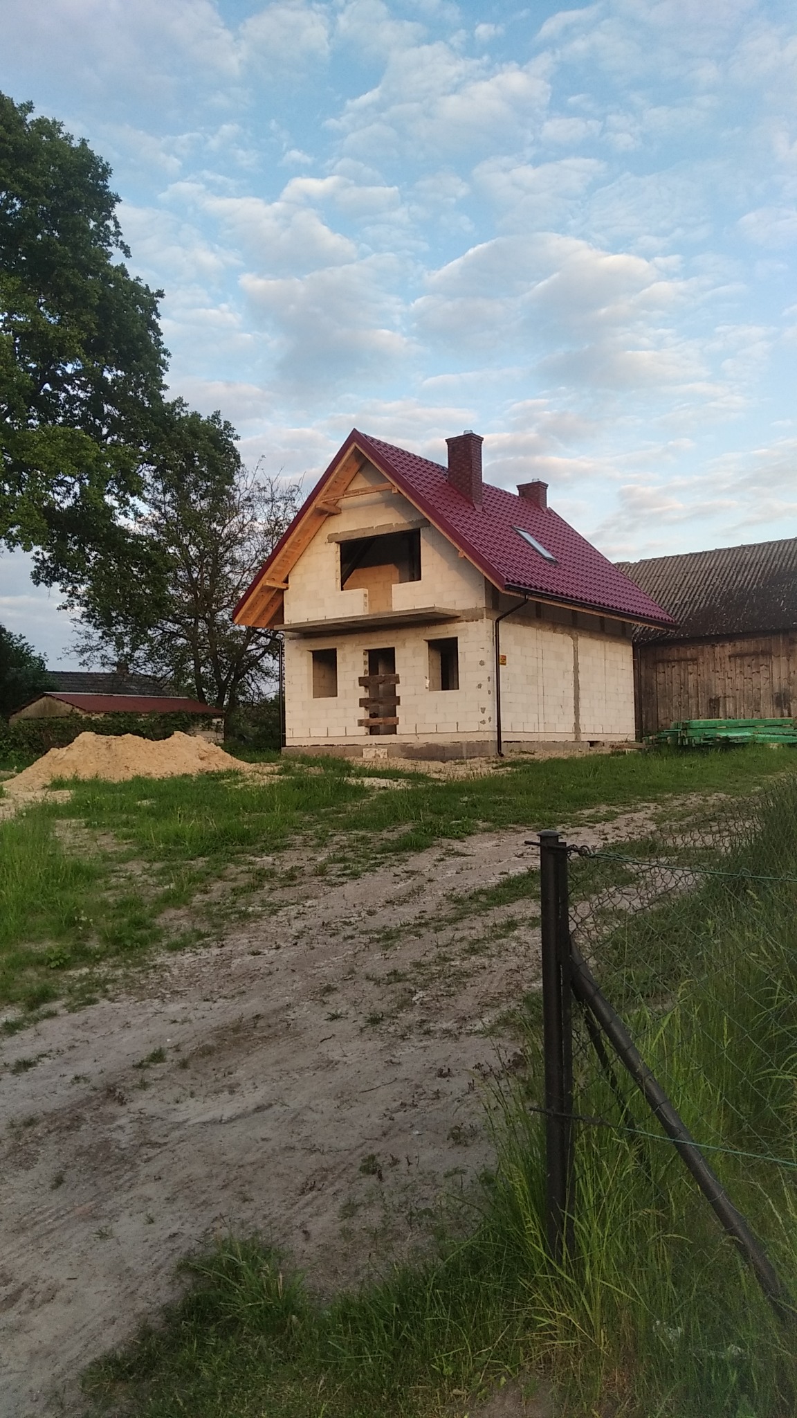 Niedokończony dom z białego pustaka z czerwoną dachówką, widoczna sterta piasku i drewniane podpory w otworach okiennych, w tle budynek gospodarczy.