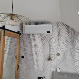 Zamontowany klimatyzator Samsung Wind Free Awant w pomieszczeniu w Prandocinie z tapetą w liście palmowe, widoczny również ozdobny żyrandol i przesuwne drzwi.