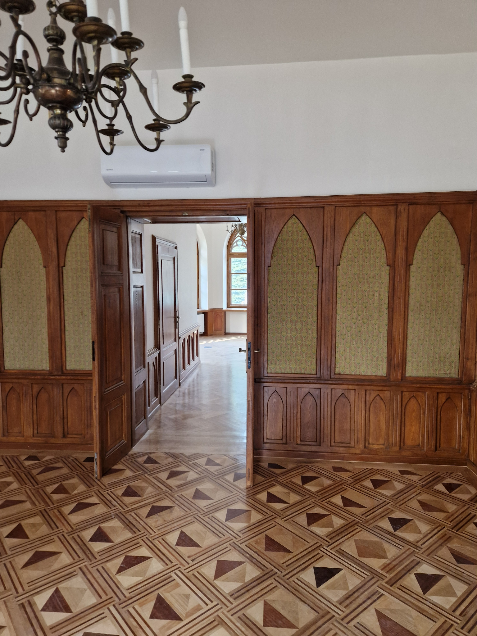 Wnętrze zabytkowego pałacu z drewnianą boazerią, parkietem w geometryczne wzory i klimatyzatorem ściennym pod żyrandolem.