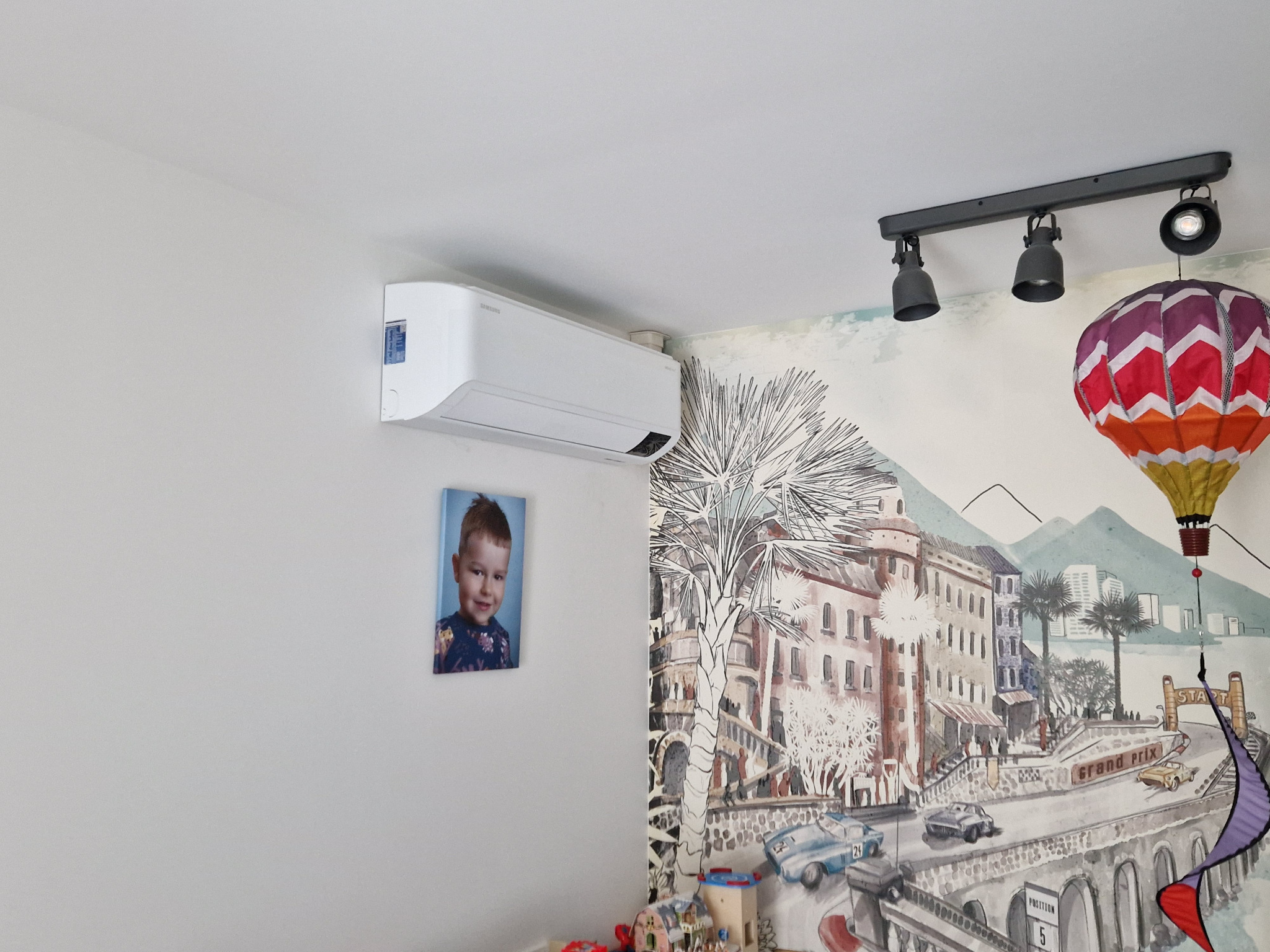 Biały klimatyzator Samsung zamontowany na ścianie w pokoju dziecięcym z muralem przedstawiającym miasto i balon na ogrzane powietrze, pod nim wisi portret chłopca.