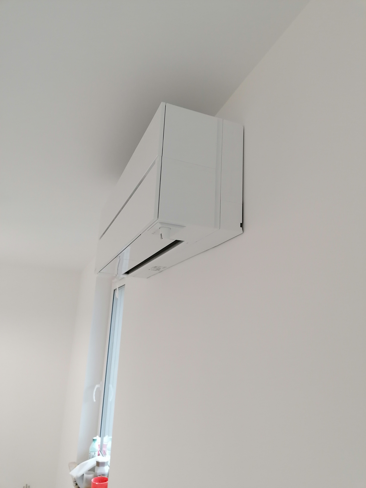 Biała klimatyzacja ścienna zamontowana w jasnym pomieszczeniu, widoczna z boku, z oknem w tle.