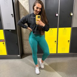 Izabela Gałęziowska Strefa Komfortu - Uśmiechnięta kobieta w sportowym stroju (czarna bluza z kapturem i turkusowe legginsy) robi selfie w szatni siłowni na tle rzędu szafek.