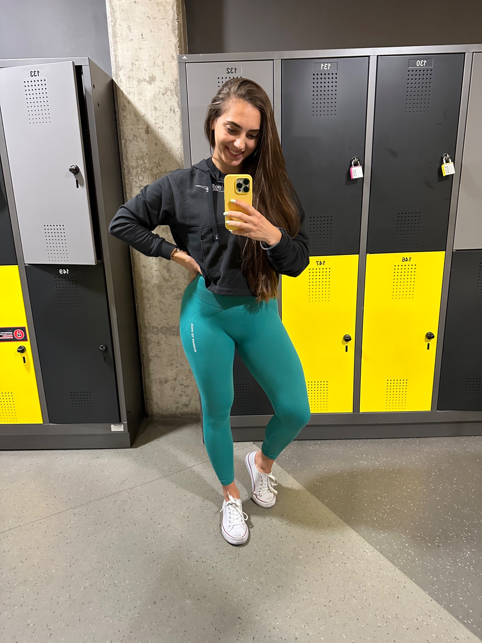 Uśmiechnięta kobieta w sportowym stroju (czarna bluza z kapturem i turkusowe legginsy) robi selfie w szatni siłowni na tle rzędu szafek.