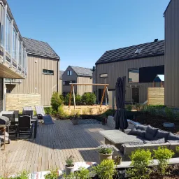 Nowoczesny taras drewniany z meblami ogrodowymi, grillem i placem zabaw w tle, otoczony budynkami o minimalistycznej architekturze.