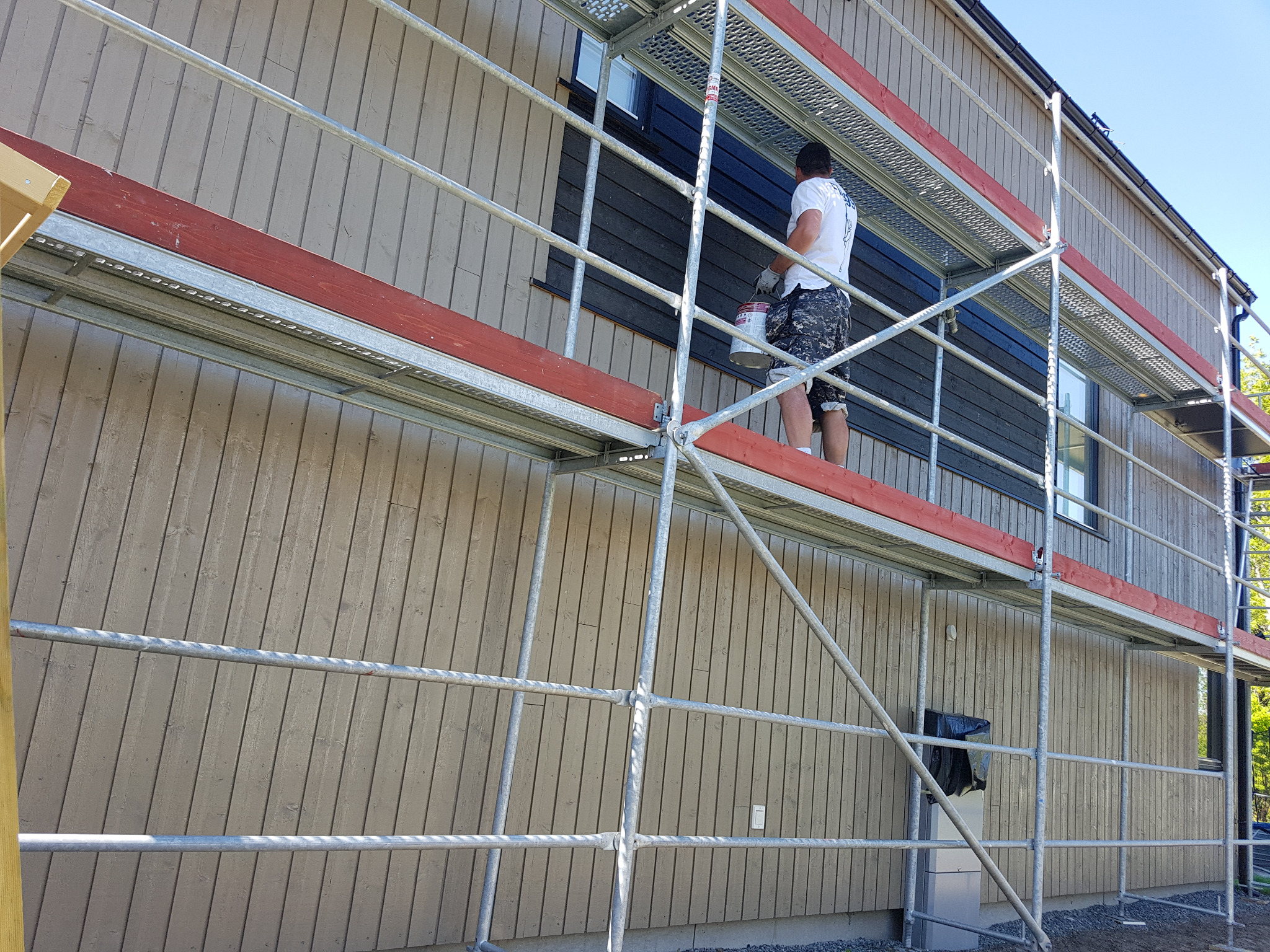 Mężczyzna maluje elewację domu z rusztowania; widoczne drewniane deski elewacyjne w szarym odcieniu oraz pomalowane na czarno obramowanie okna.