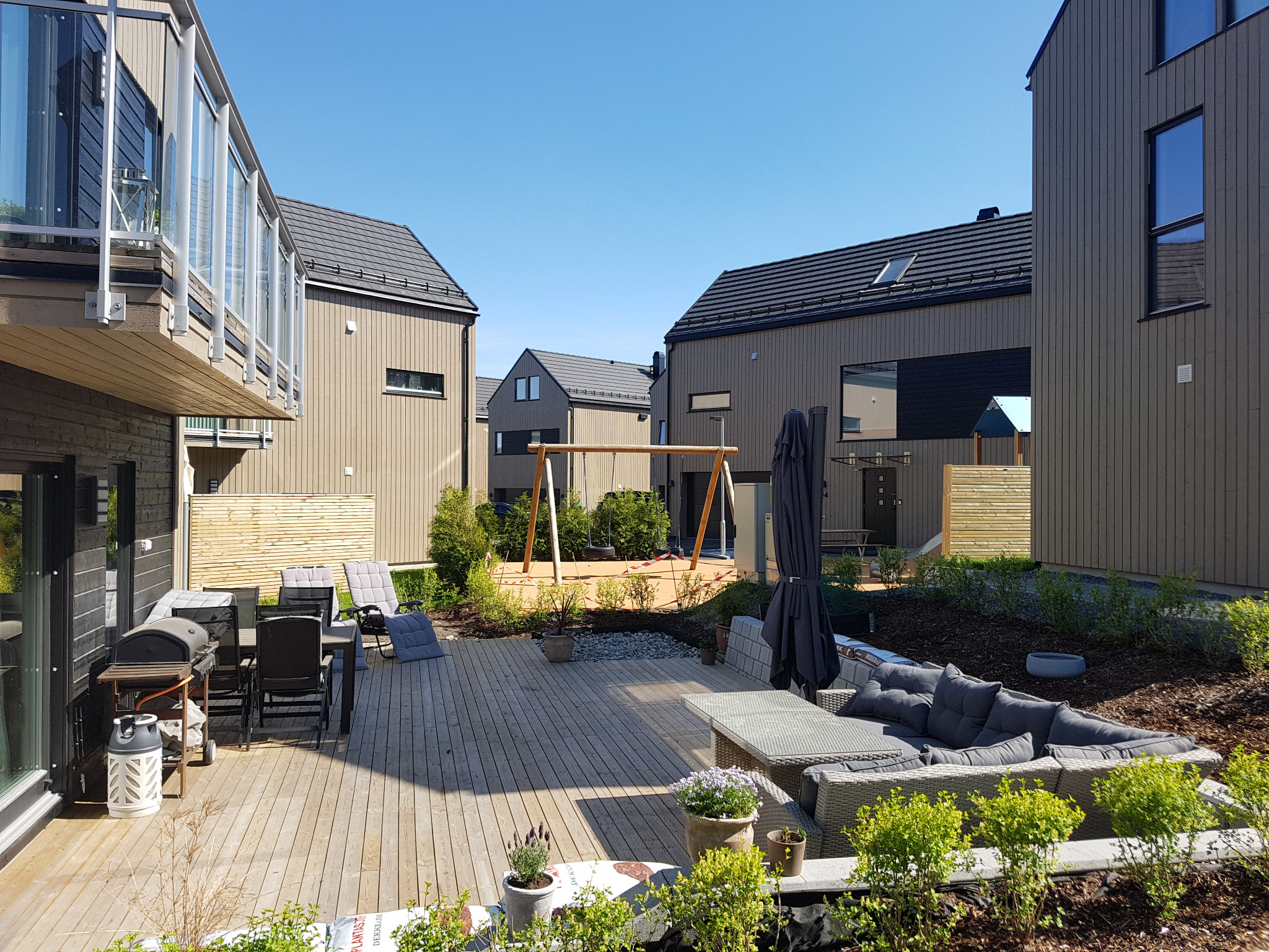 Nowoczesny taras drewniany z meblami ogrodowymi, grillem i placem zabaw w tle, otoczony budynkami o minimalistycznej architekturze.