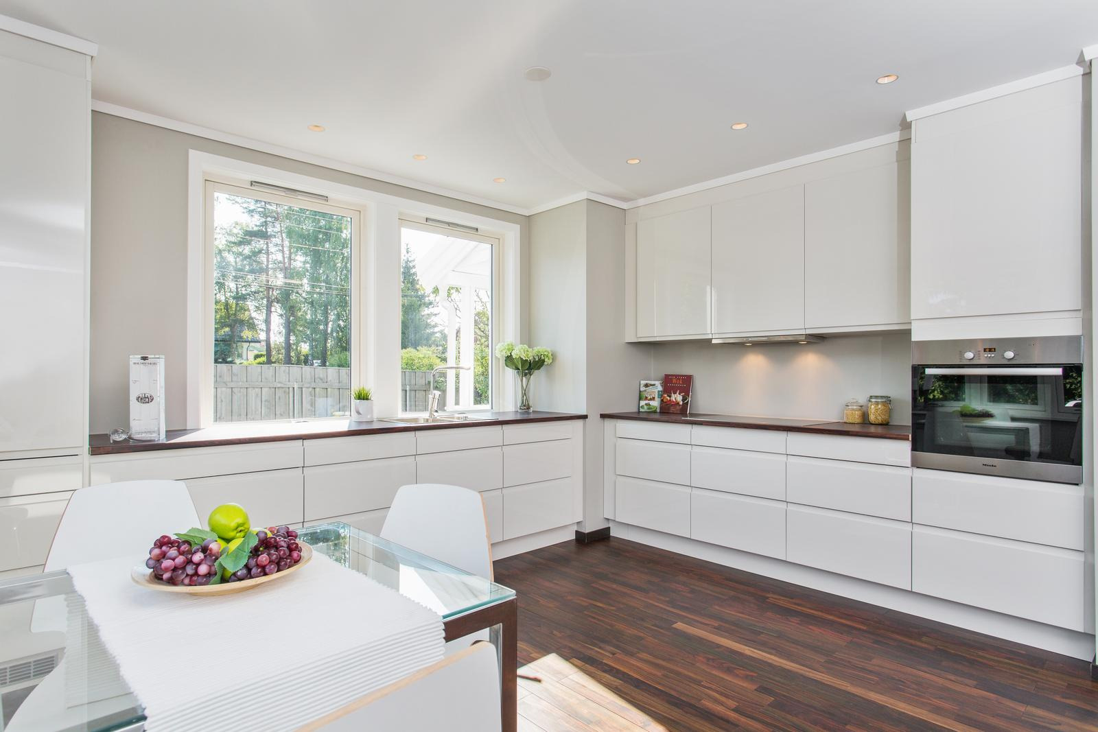 Jasna, minimalistyczna kuchnia z białymi, lakierowanymi frontami szafek, drewnianym blatem i podłogą, oraz szklanym stołem z owocami w tle okna.