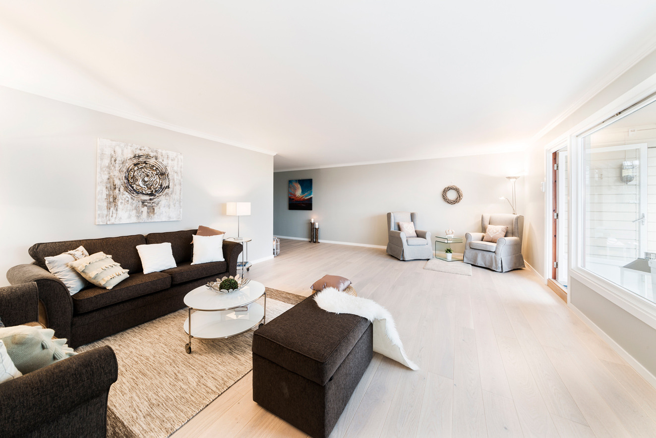 Jasne, przestronne wnętrze salonu z drewnianą podłogą, ciemną sofą z poduszkami, okrągłym stolikiem kawowym na kółkach, pufą z futerkiem, dwoma fotelami w stylu angielskim i obrazami na ścianach.