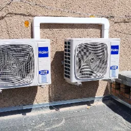 Dwa klimatyzatory Haier zamontowane na zewnątrz budynku, połączone białym przewodem, widoczne okno i fragment muru z cegły.