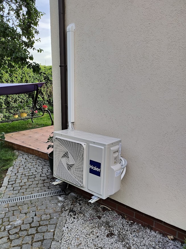 Zewnętrzna jednostka klimatyzacji Haier zamontowana na elewacji domu, z widocznym odprowadzeniem skroplin i kostką brukową wokół.